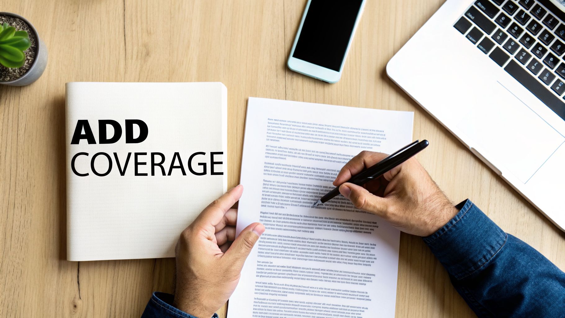 Overhead shot of a person signing a document, with a notebook displaying 'ADD COVERAGE' on a wooden desk.